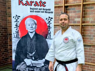 Auf dem Bild ist unser Spartenleiter Belal Chahrour zu sehen, wie er mit guter Laune vor dem Banner des Karate Vereins Budokan Göttingen e.V. steht.